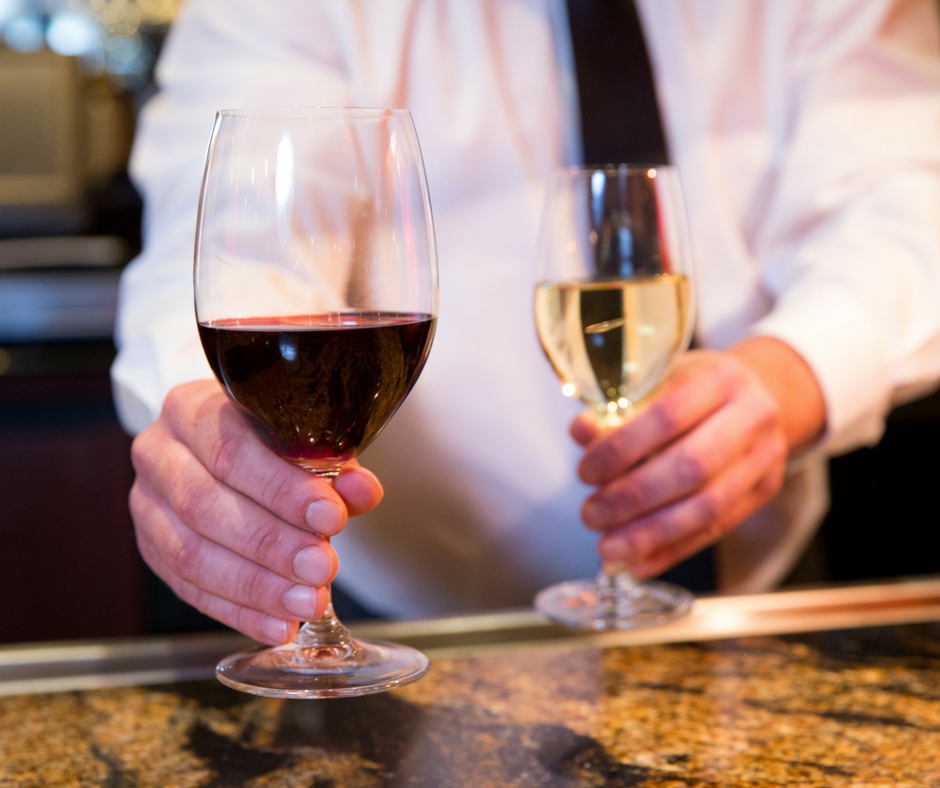 Two glasses of wine, one red and one white, being held out by a bartender in a white shirt with a black tie over a marble bar